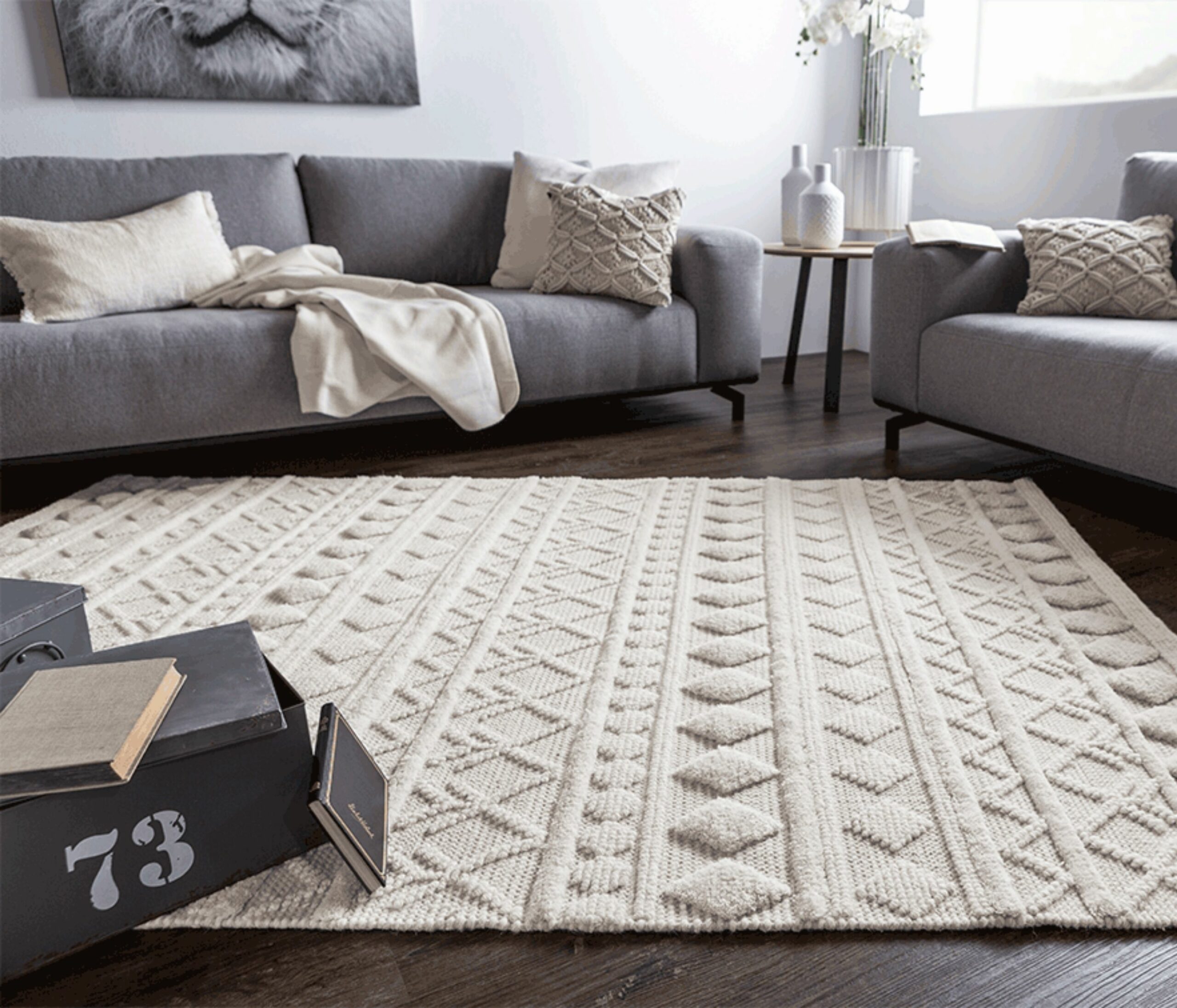 Bequemer Teppich in Creme mit Strukturmuster im Wohnzimmer mit grauen Sofas und dunklem Boden in Holzoptik.